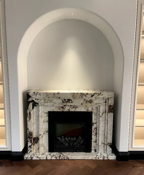 White Volakas marble fireplace surround, with stepped tiers and bold veining, in a furnished room