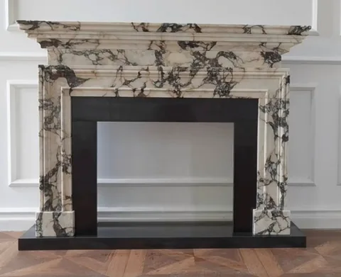White Volakas marble fireplace surround, with stepped tiers and bold veining, in a furnished room