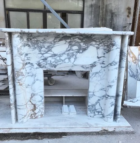 White Volakas marble fireplace surround, with a straight recessed opening and bold veining, in a workshop