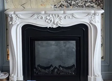 White Volakas marble fireplace surround, with carved scrollwork and a plain surface, in a furnished room